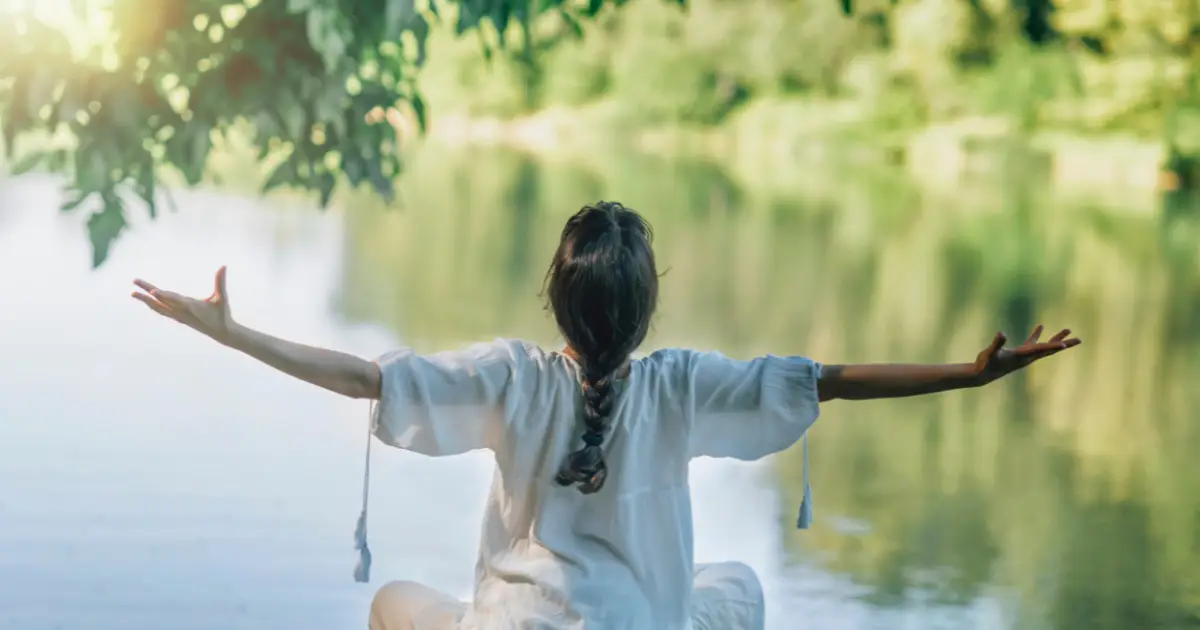 Woman practicing mindfulness meditation by a lake, sitting with arms outstretched.