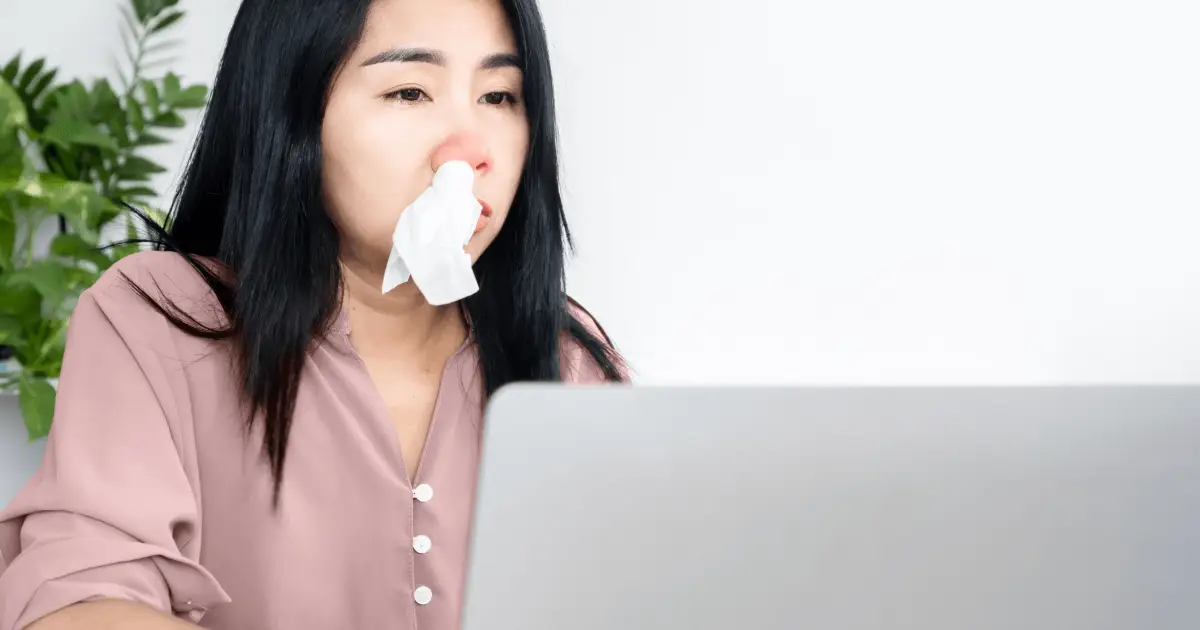 Woman with tissue in mouth, sitting at laptop, likely due to allergic rhinitis.