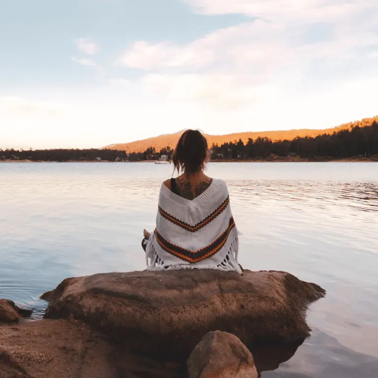 A woman peacefully seated on a rock, using a grounding exercise to reduce stress near a serene lake