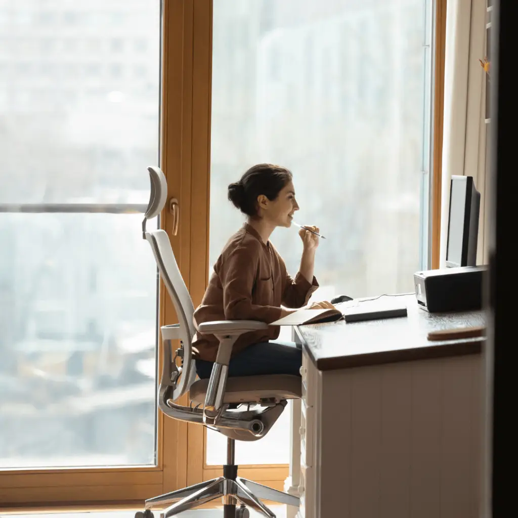 Woman seated at a desk by a window, utilizing ergonomic furniture for posture correction.