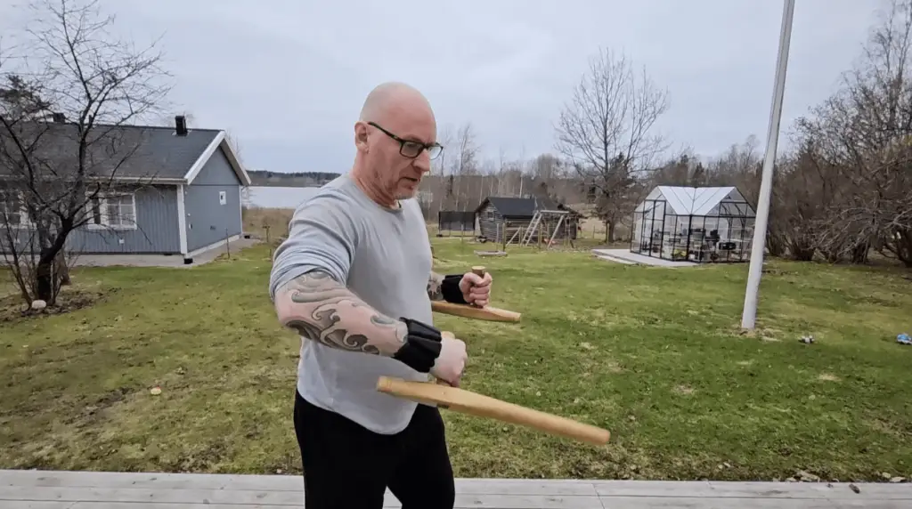A man showcasing a tattoo on his arm holds a wooden bat, accompanied by black wrist and ankle weights.