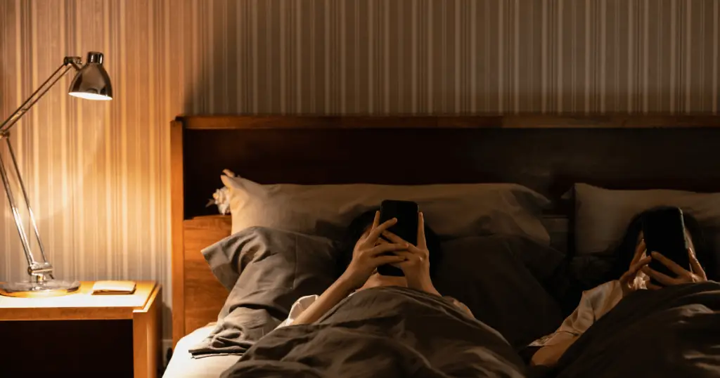 Two people laying in bed, holding phones and smiling.