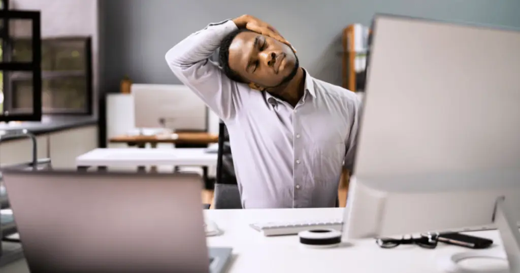 A man practices desk yoga, sitting at his desk with his head in his hands, seeking relief from stress and tension.