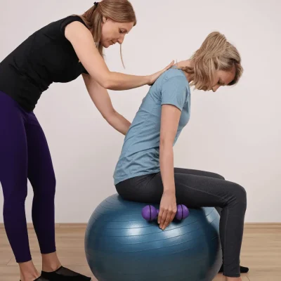 A woman engages in a physical therapy exercise on an exercise ball, guided by another woman. Professional assistance is recommended for posture correction