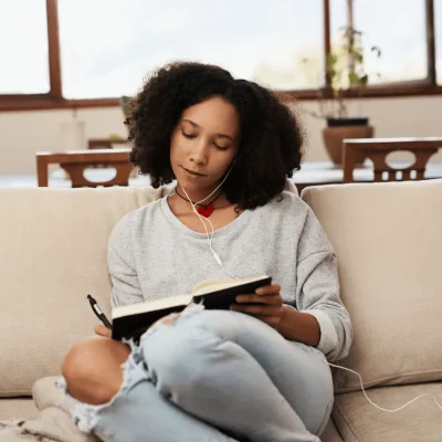A young woman engrossed in a book, sitting on a home couch. She is focused on shadow work and working on journals.